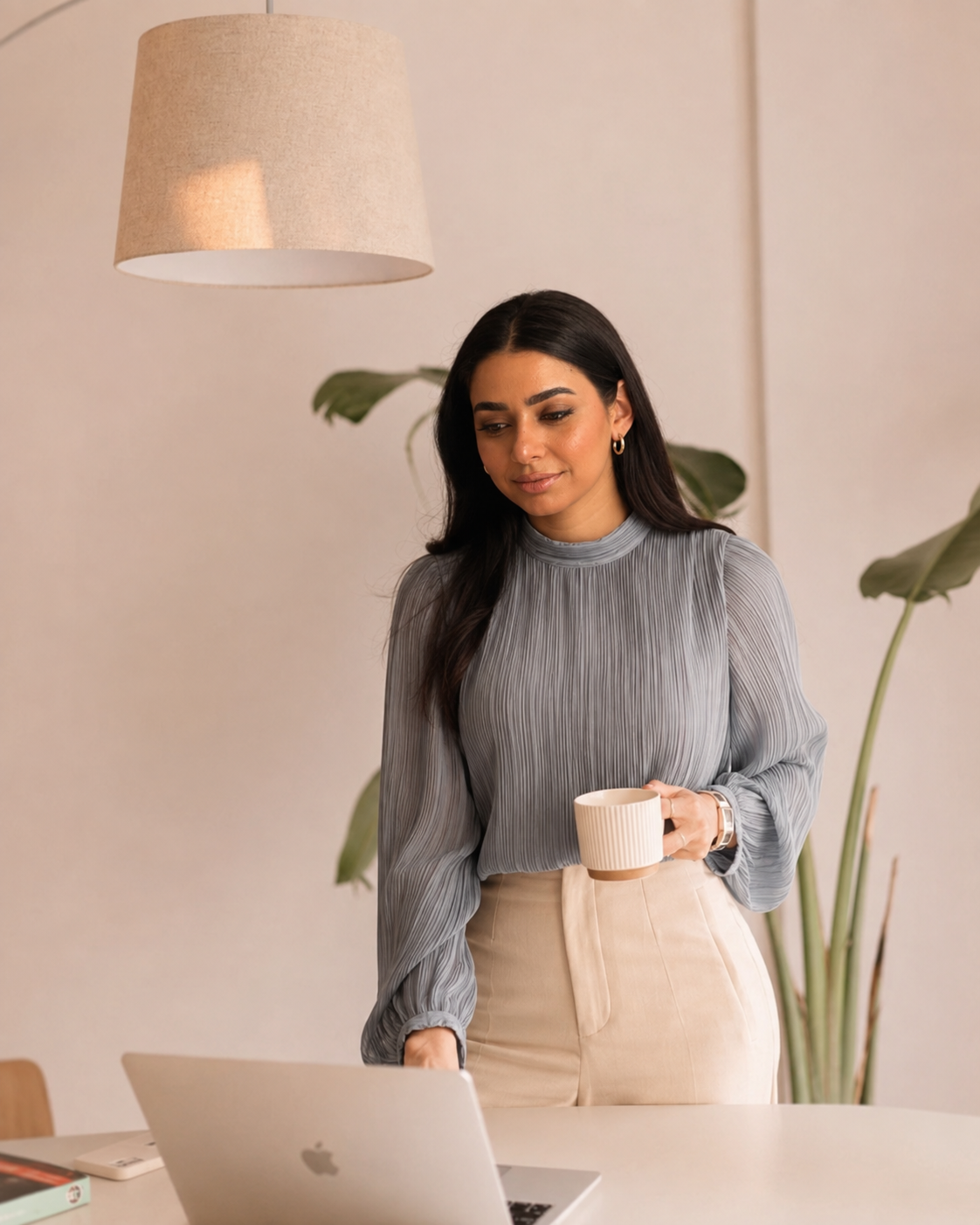 Amel Amamou standing at a desk with a laptop and a cup of coffee in a calm, light-filled workspace.